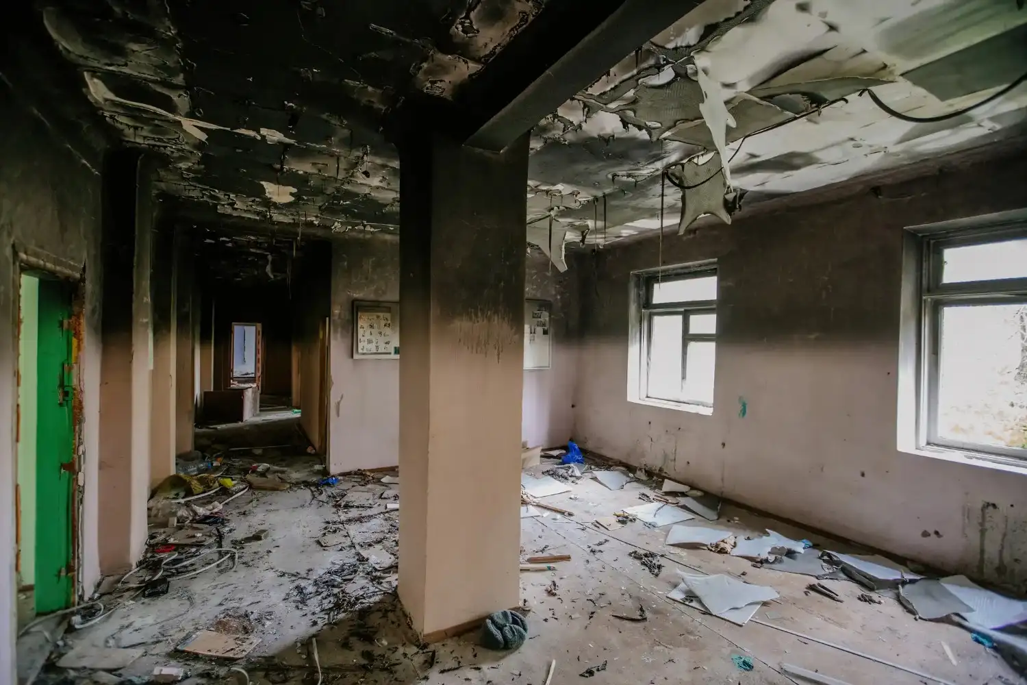 An interior view of a fire-damaged building shows a hallway with peeling paint, a damaged ceiling, and debris scattered on the floor.