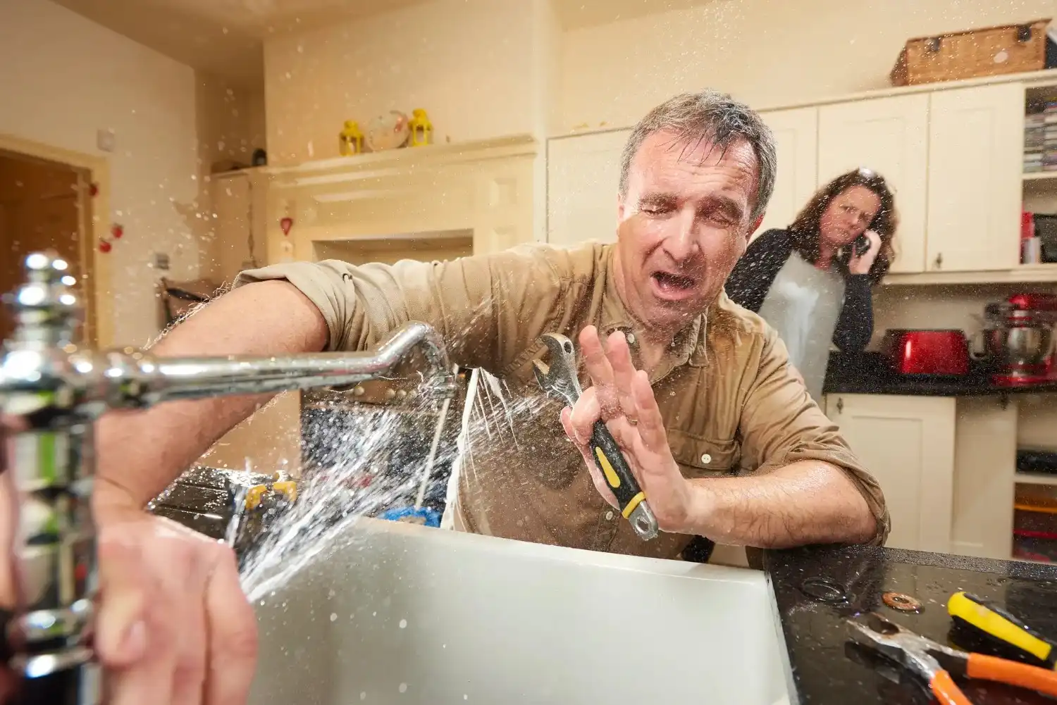 A man attempting to stop a water leak from a kitchen faucet with his hand, while a woman calls on a cell phone in the background