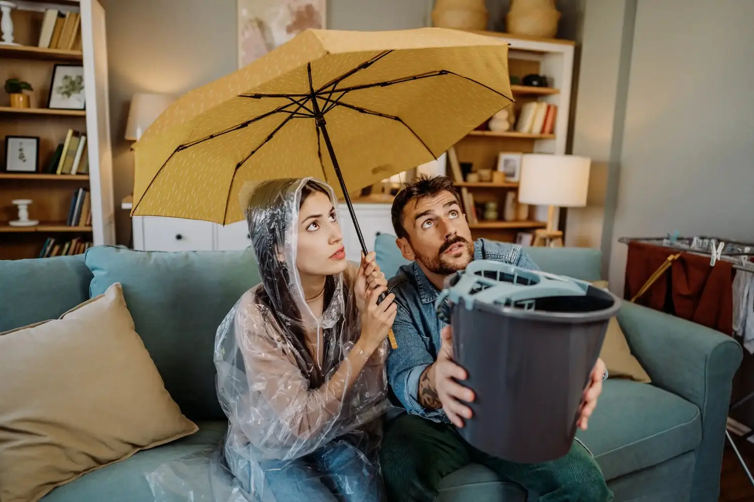 A couple sitting on a couch under an open umbrella, with the man holding a bucket to catch water from a leaky ceiling