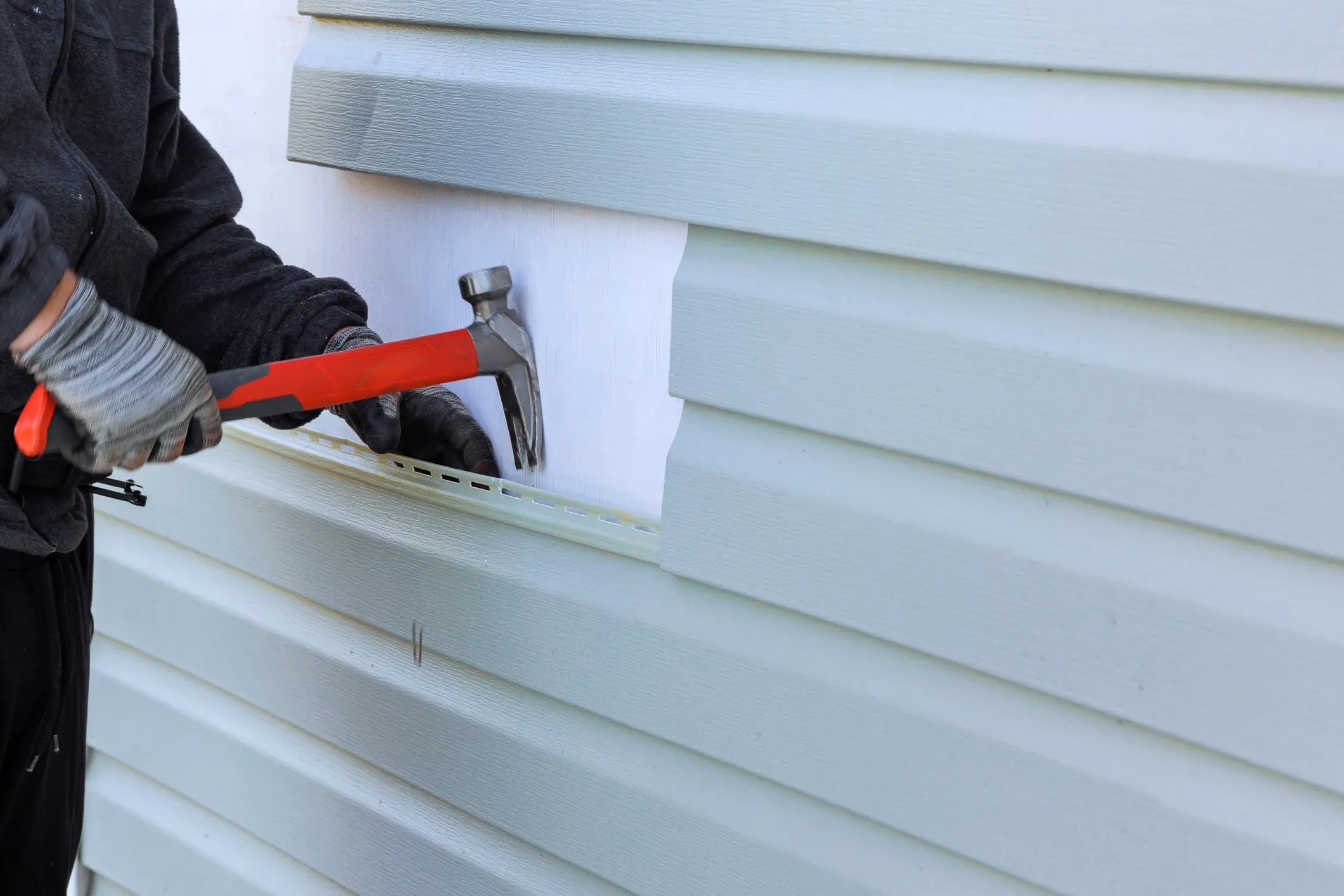 Worker installing vinyl siding with hammer and protective gloves