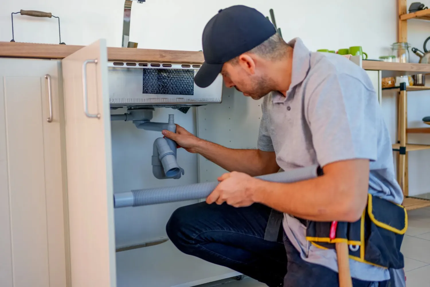 Plumber in cap inspecting under-sink pipes and plumbing connections