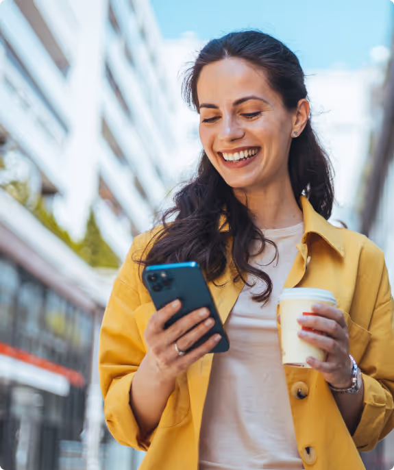 Smiling woman in yellow jacket holding a smartphone and a takeaway coffee cup outdoors in an urban setting.