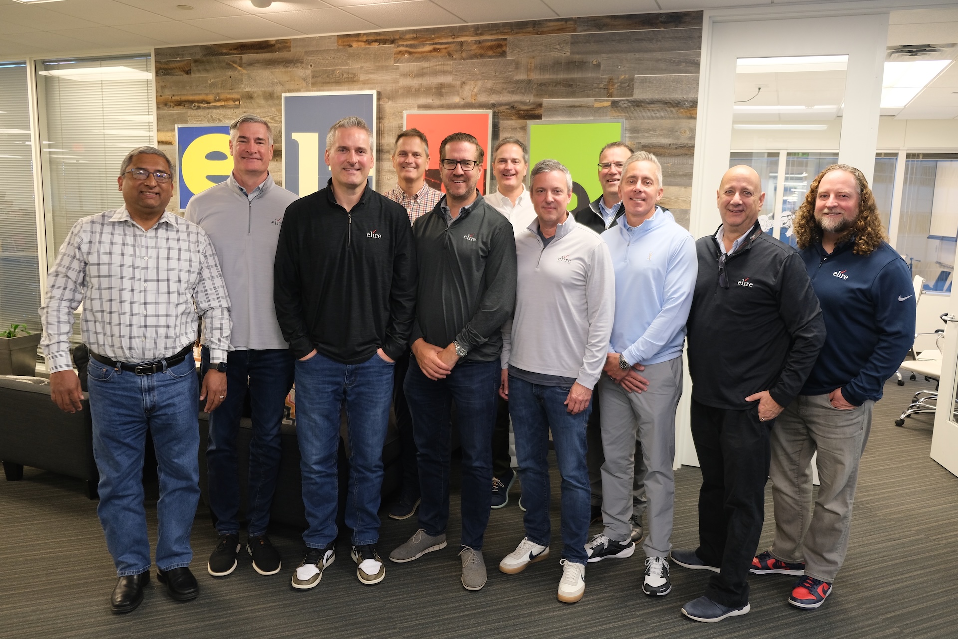 Group of eleven men standing indoors in casual attire, smiling at the camera with colorful framed letters on a wooden wall behind them.