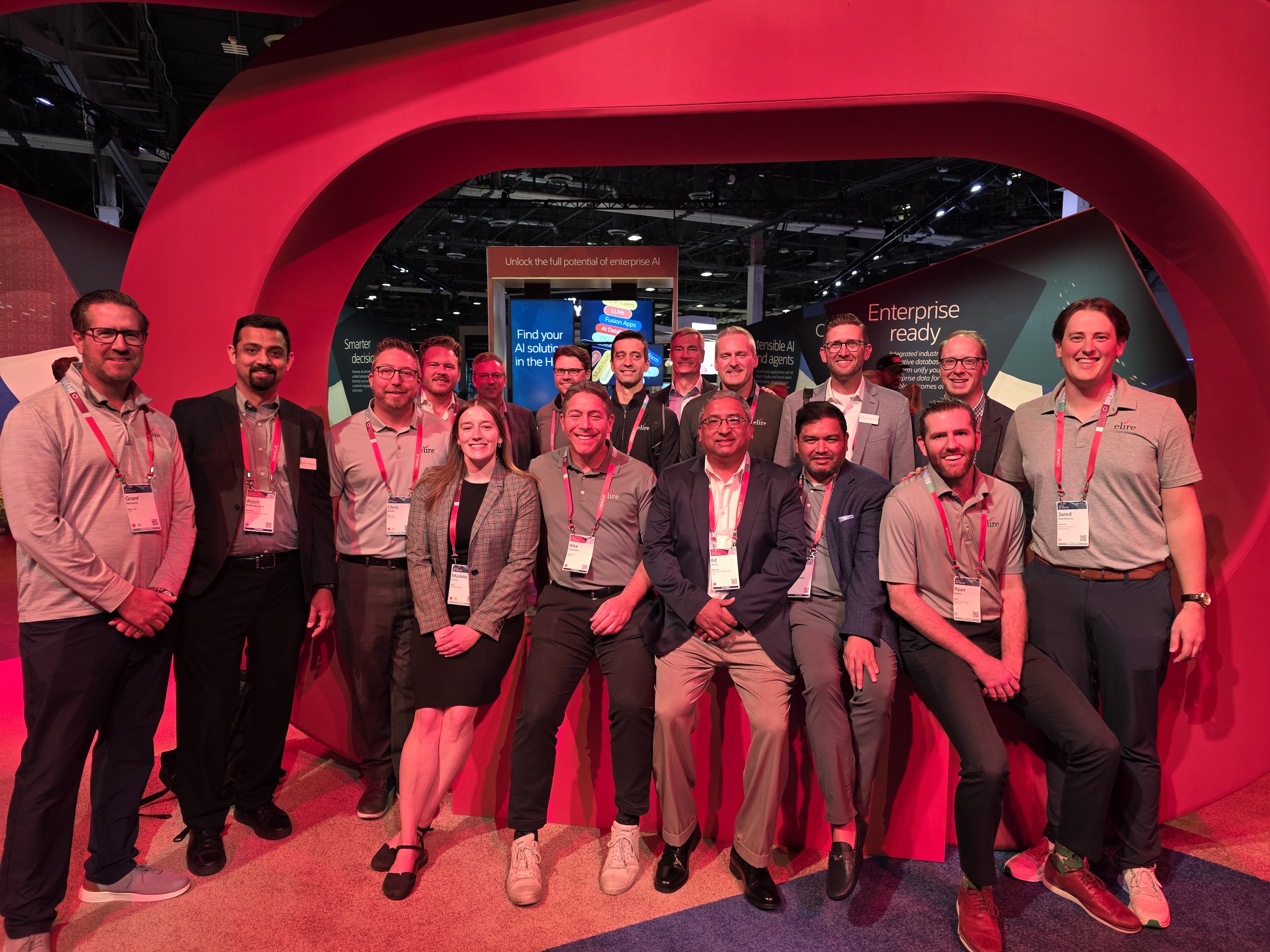 Group of 16 business professionals posing together under a large red arch at a tech event with AI-related displays in the background.