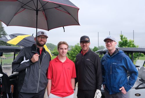 Four men standing outdoors on a rainy day at a golf course, with two holding umbrellas and golf carts in the background.