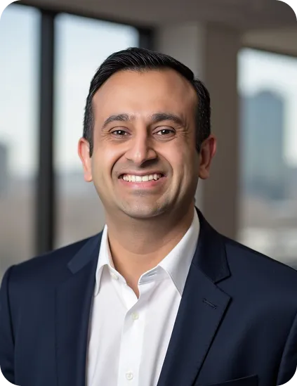 Smiling man wearing a dark blazer and white shirt with blurred office windows in background.