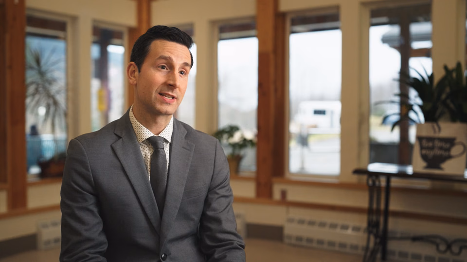 Man in a gray suit speaking while seated in a room with large windows and plants.