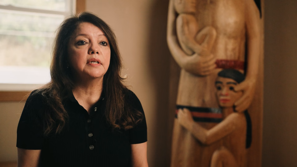 Woman with long dark hair wearing a black shirt sitting indoors next to a wooden sculpture of two figures.