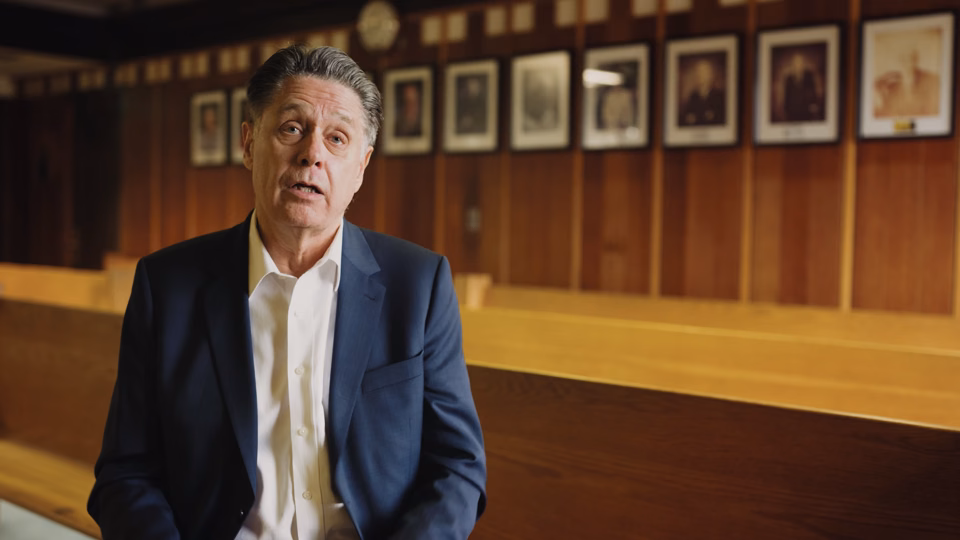 Middle-aged man in a navy blazer and white shirt sitting indoors with wooden benches and framed pictures on the wall behind him.