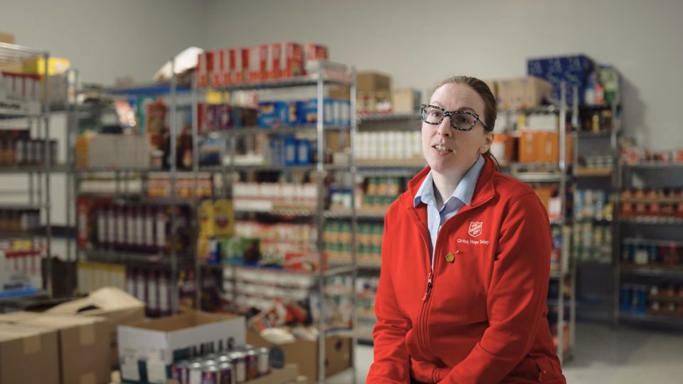 Woman in a red Salvation Army jacket sitting in front of shelves stocked with food and supplies in a storage or distribution center.