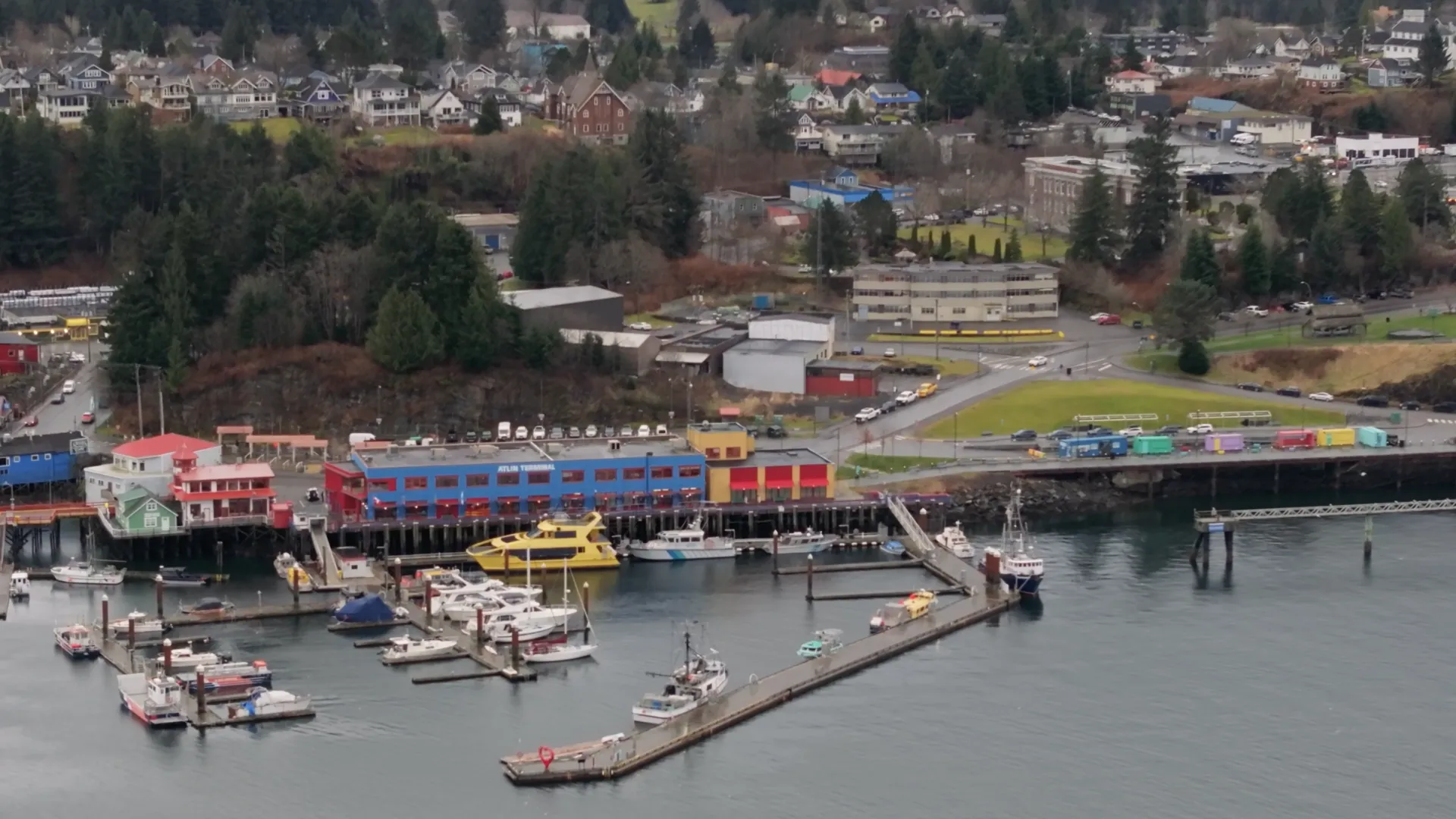 Aerial view of a marina with multiple boats docked, colorful buildings including a blue and red terminal, and a town with houses and trees in the background.