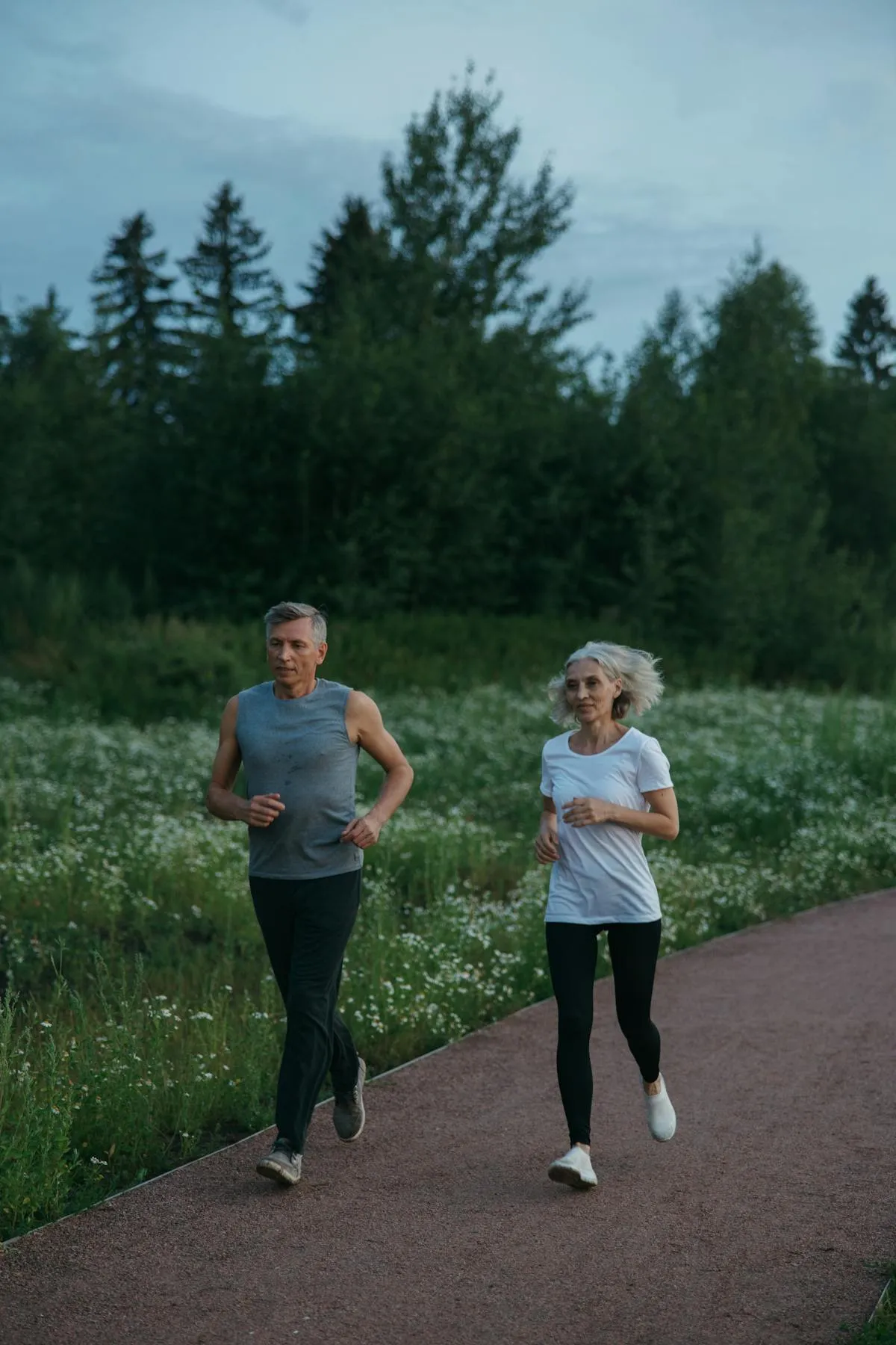 Mature man and woman jogging side by side on a pathway surrounded by greenery and wildflowers.