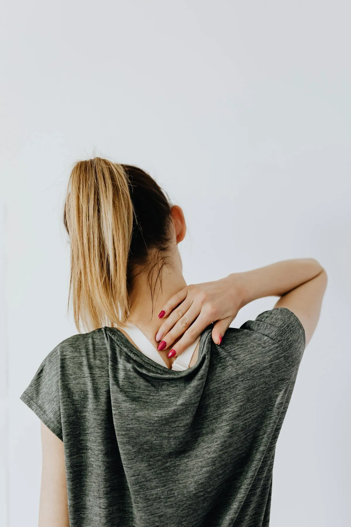 Woman with blonde ponytail wearing a gray shirt, holding the back of her neck as if experiencing neck pain.