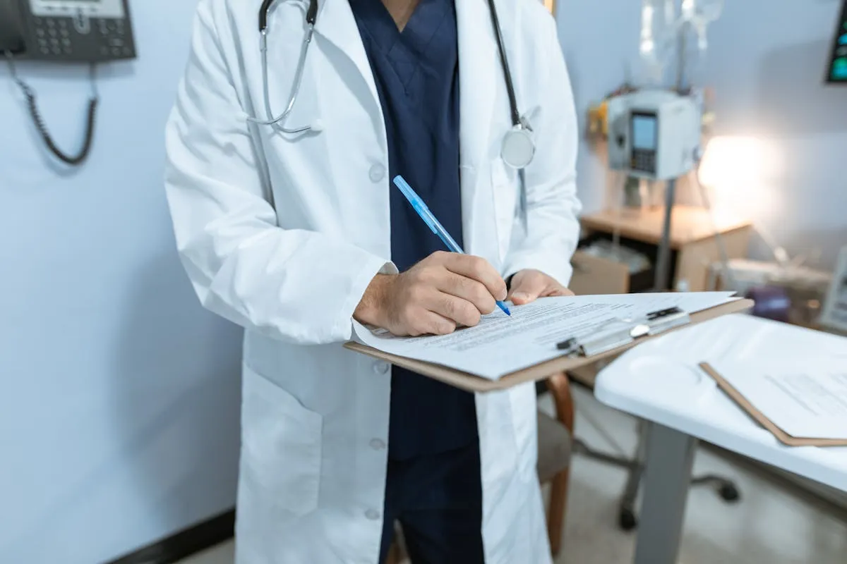 Doctor in white coat writing on a clipboard in a medical office with stethoscope around neck.