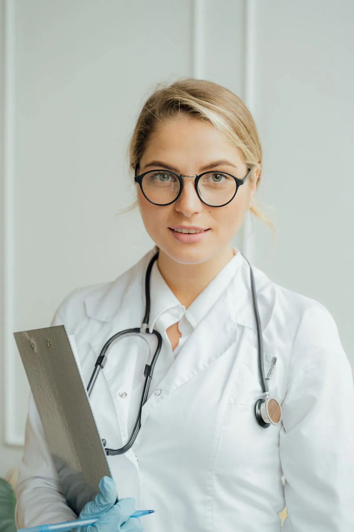 Female doctor with glasses, white coat, stethoscope, and clipboard looking at the camera.