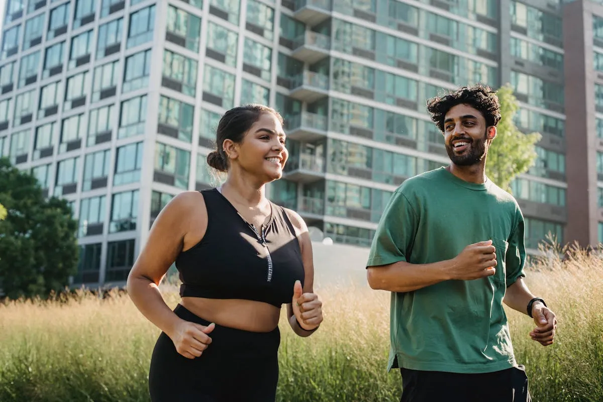 Smiling couple jogging outdoors near tall urban buildings and greenery on a sunny day.
