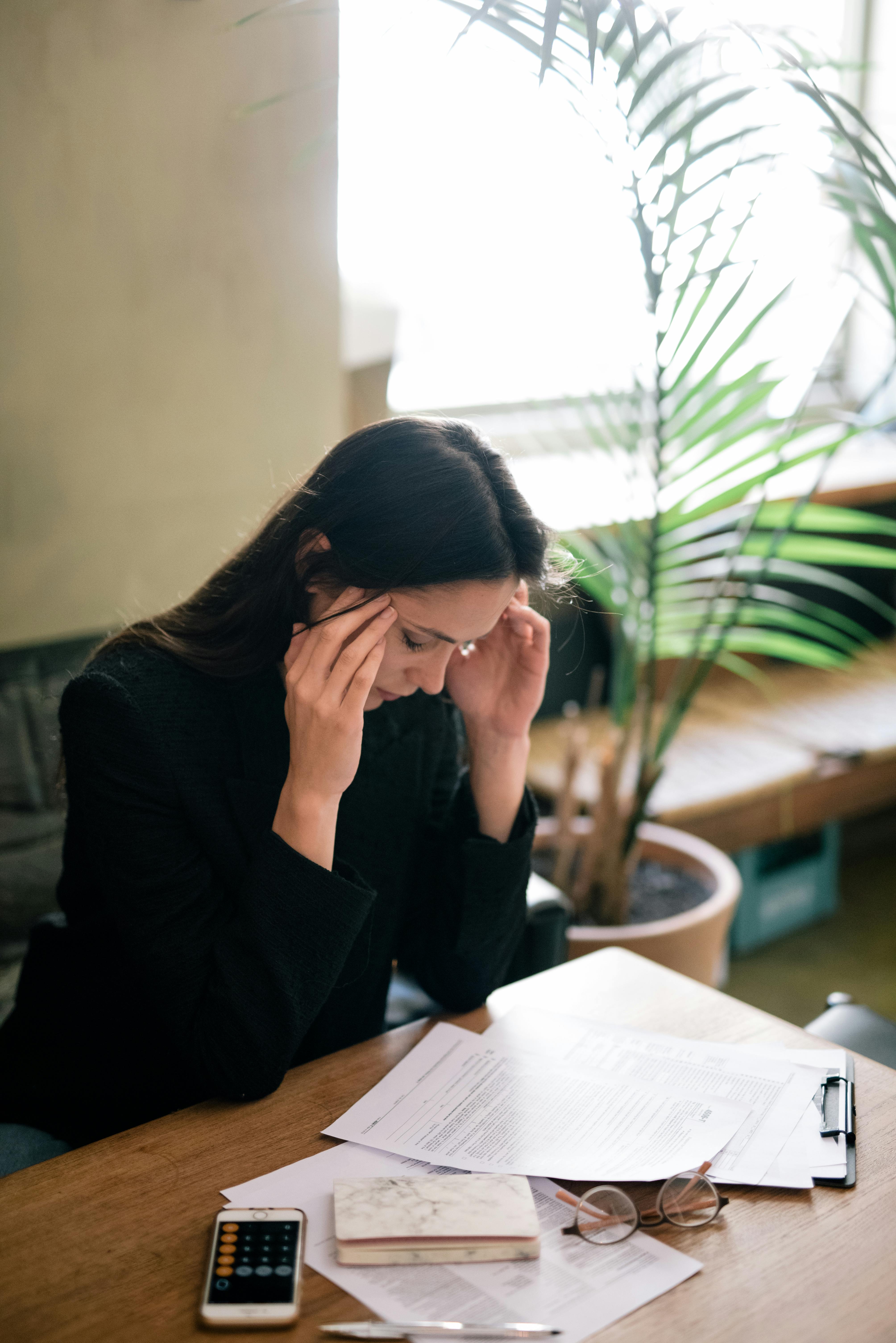 Woman sitting at a desk holding her temples, appearing to have a headache, with papers, glasses, a phone, and a notebook on the table.
