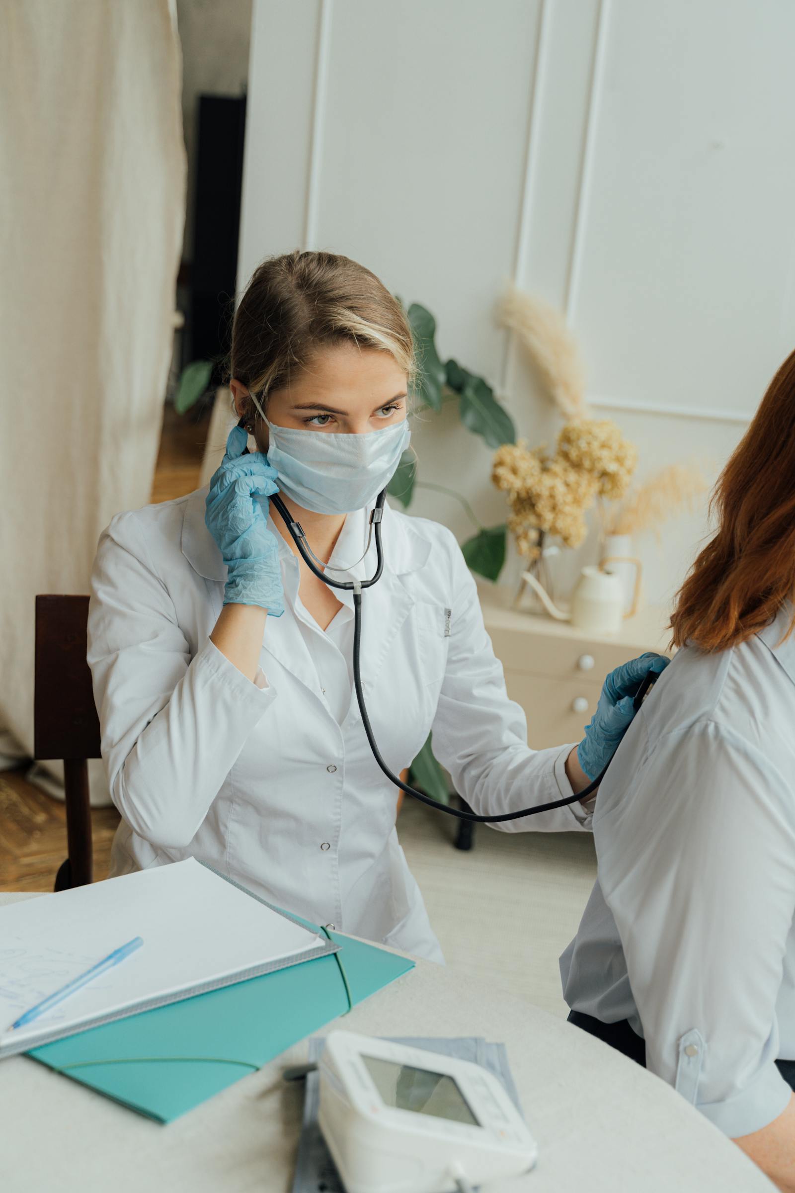 Female doctor wearing a mask and gloves using a stethoscope to examine a seated patient.