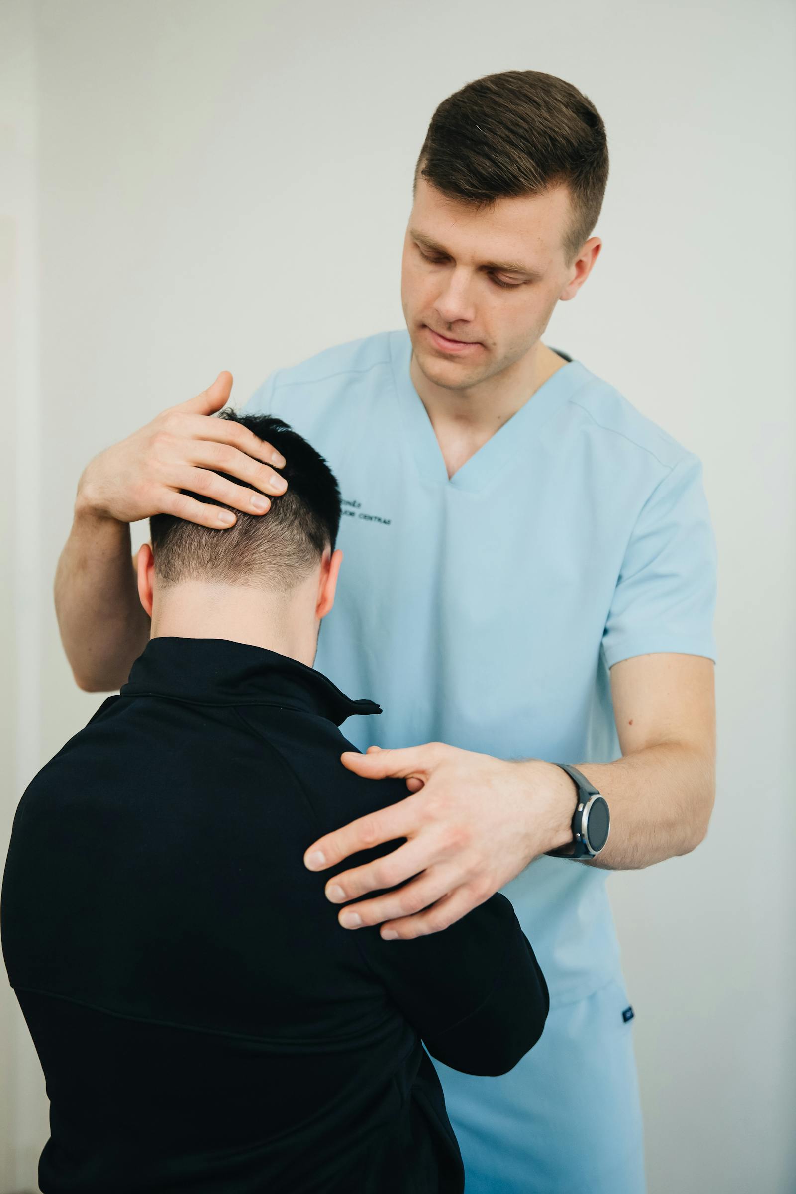 Physiotherapist in light blue scrubs gently examining a patient's neck and shoulder.