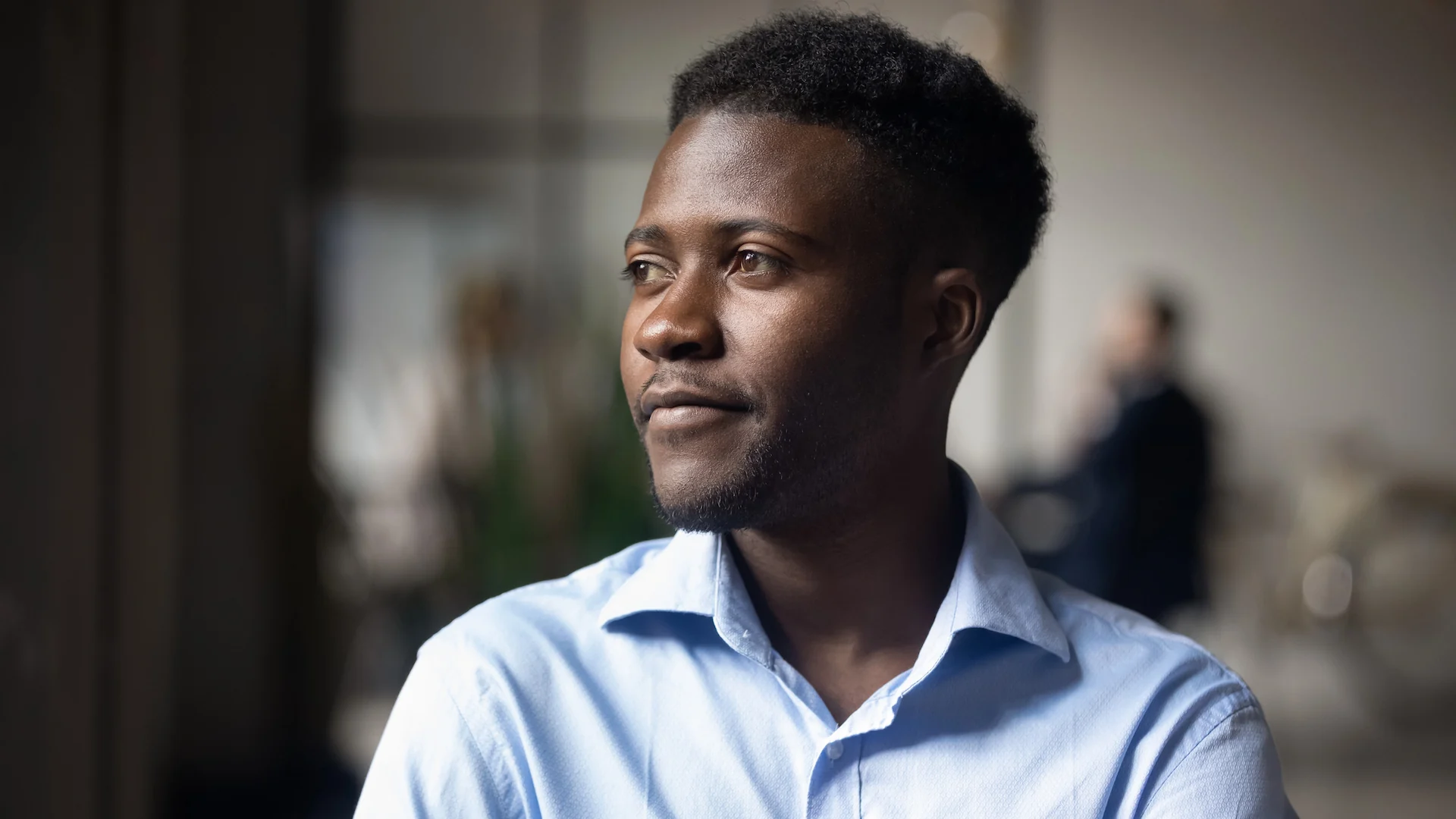A young Black man in a light blue shirt looking thoughtfully off-camera in an office setting.