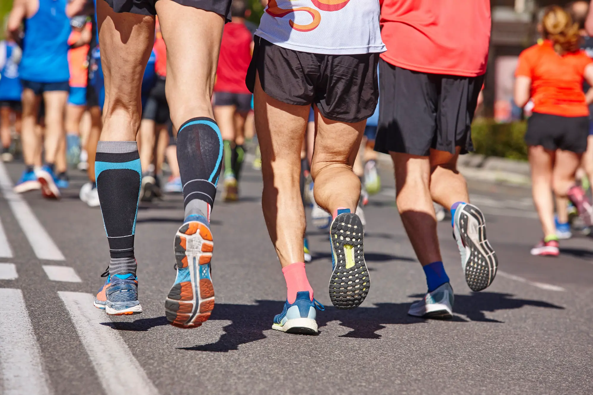 Runners' legs moving forward on an asphalt road during a marathon, showing athletic wear, compression socks, and various running shoe treads.