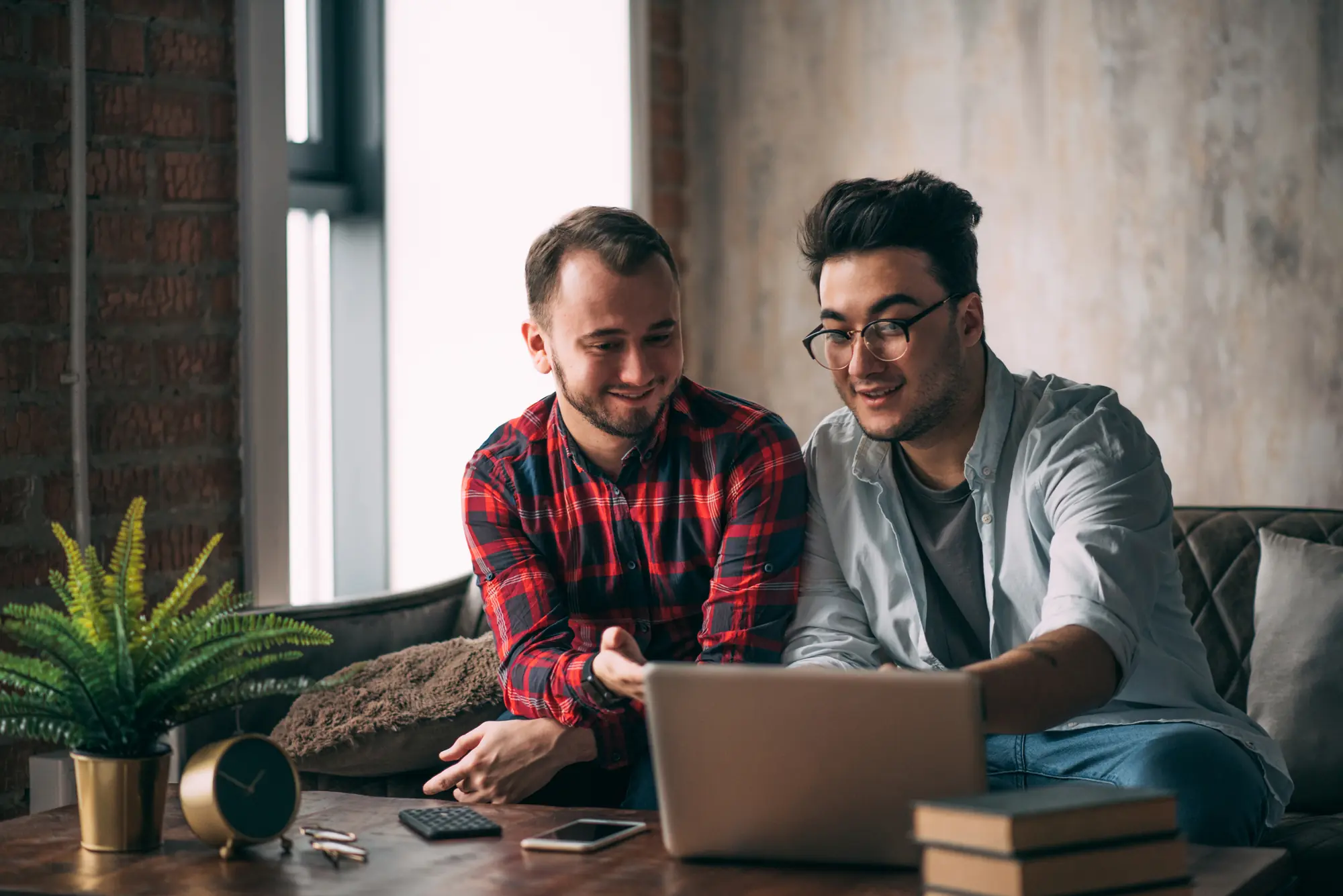Two friends or colleagues working on a laptop in a cozy, loft-style room with brick and textured walls.