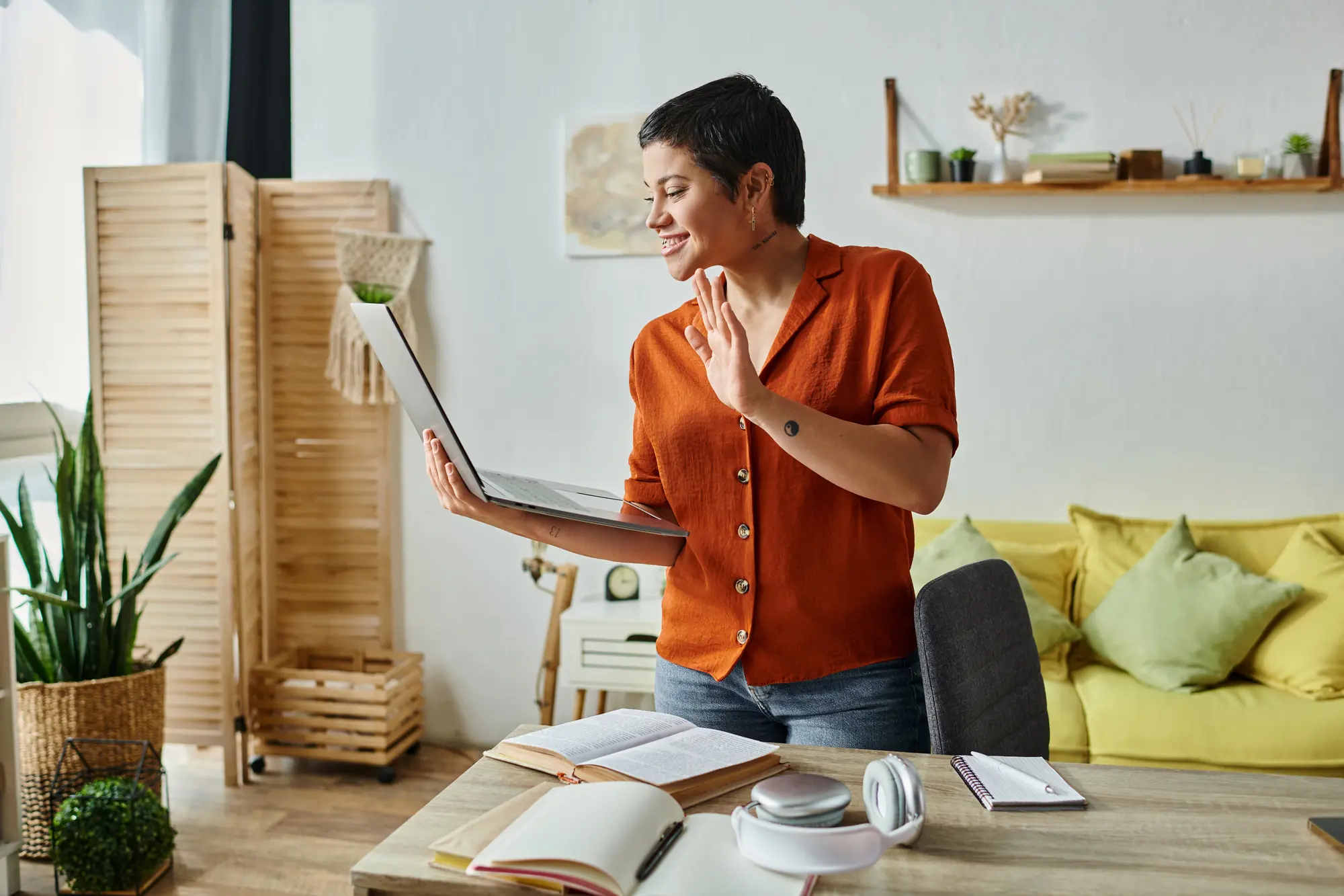 A woman with short hair waving at a laptop during a video call in her home office.