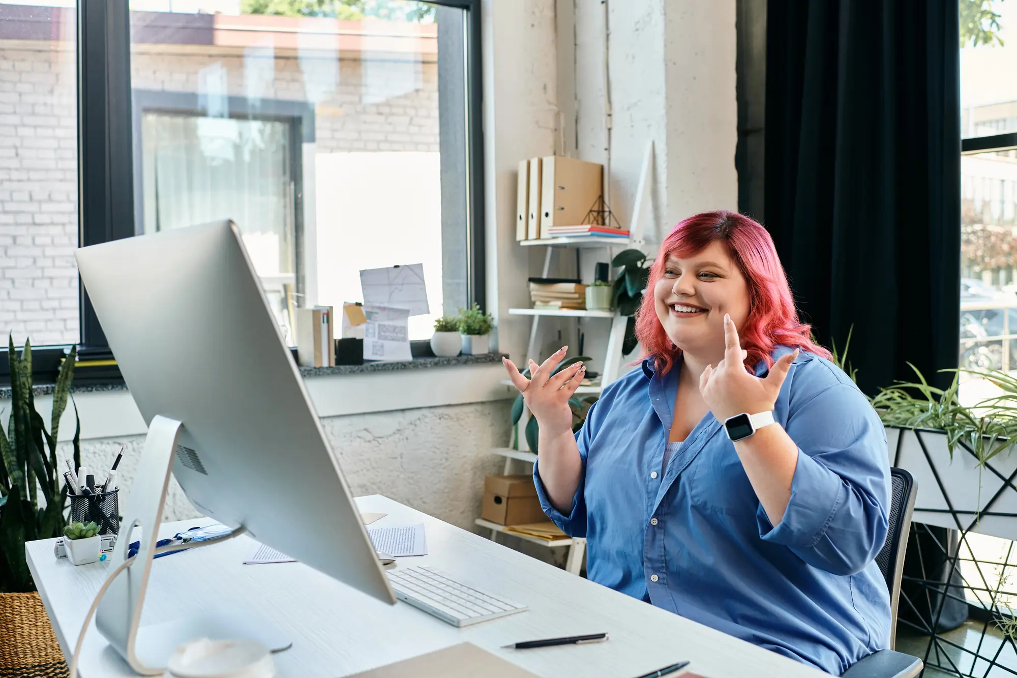 A smiling woman with pink hair sitting at a desk and talking on a video call.