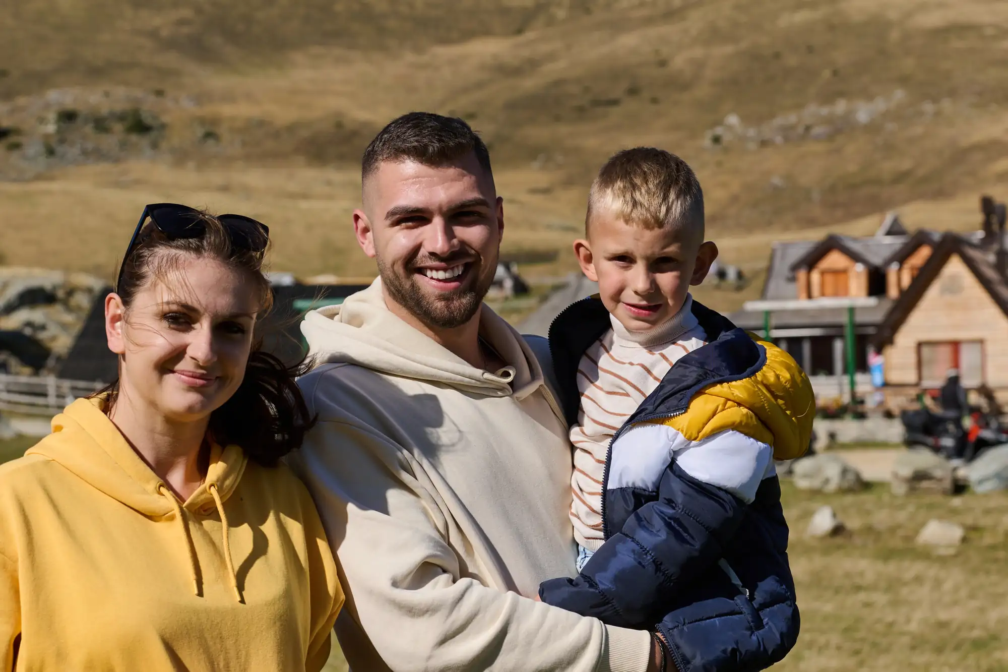 A smiling family of three posing outdoors in a mountainous, grassy landscape with cabins behind them.

