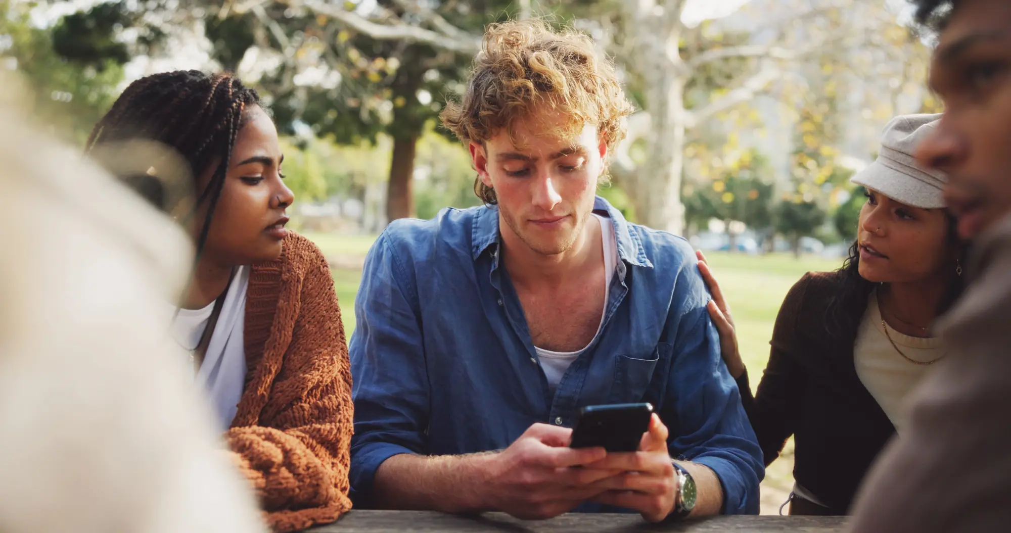 A young man looking at his phone while friends comfort him at an outdoor table.


