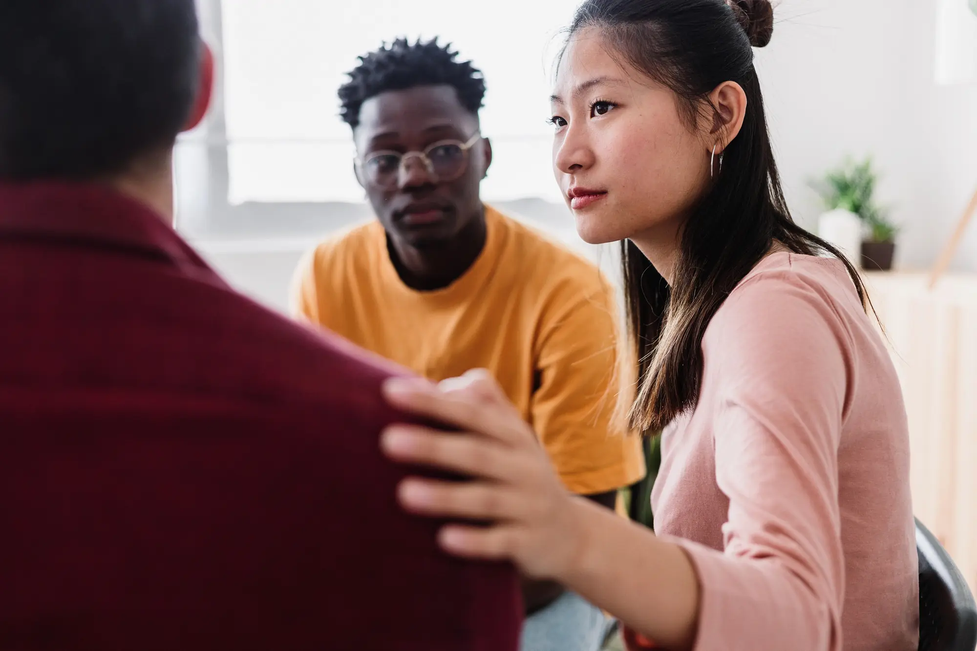 Diverse group of young adults in a support setting, with one woman placing a reassuring hand on a person's shoulder.


