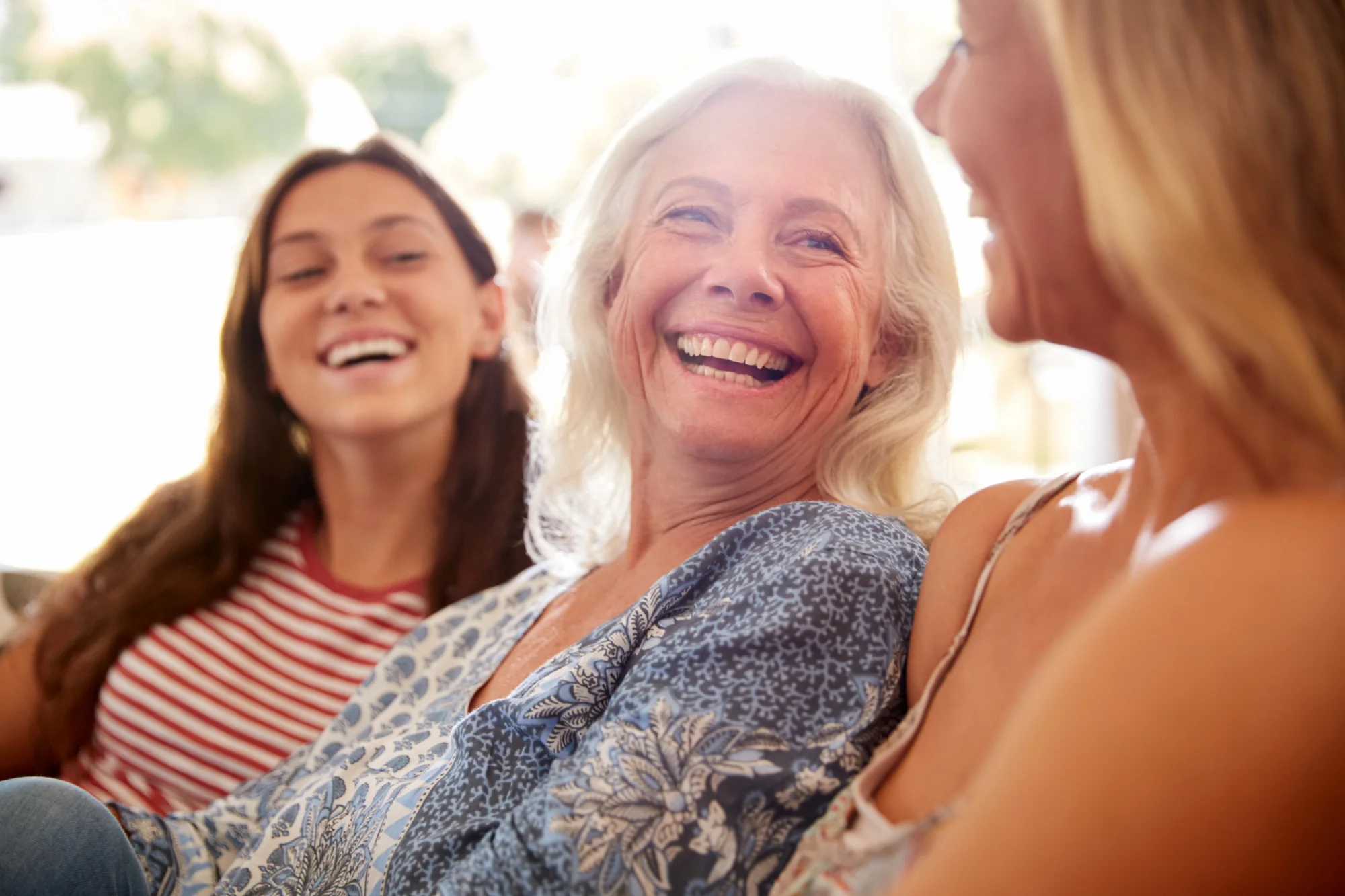 Three women of different generations laughing together in a bright, relaxed setting.


