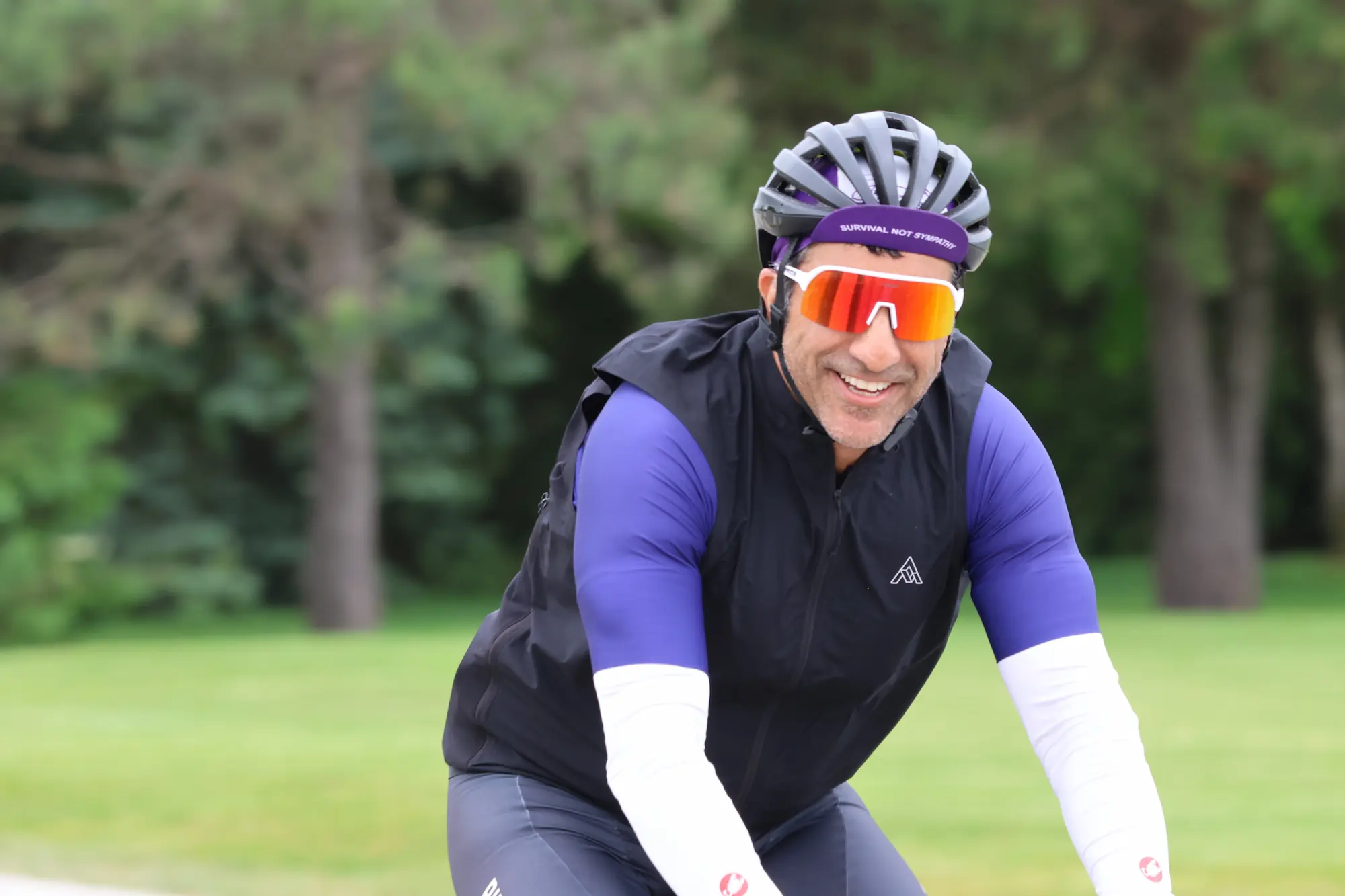 A man in cycling gear, wearing a helmet and bright sunglasses, smiling on his bike outdoors.