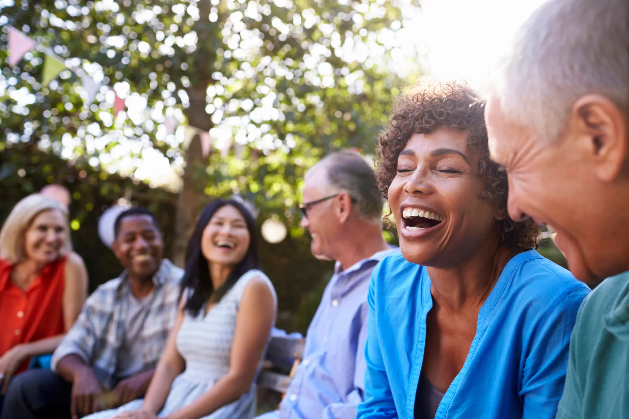 Diverse group of middle-aged and older friends laughing together outdoors at a sunny party.

