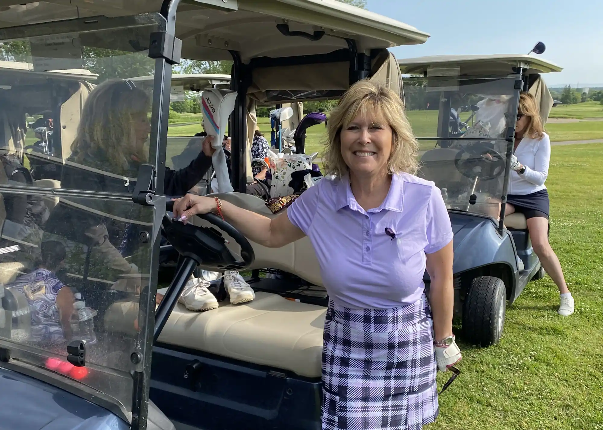 A woman smiling next to a golf cart on a sunny golf course.

