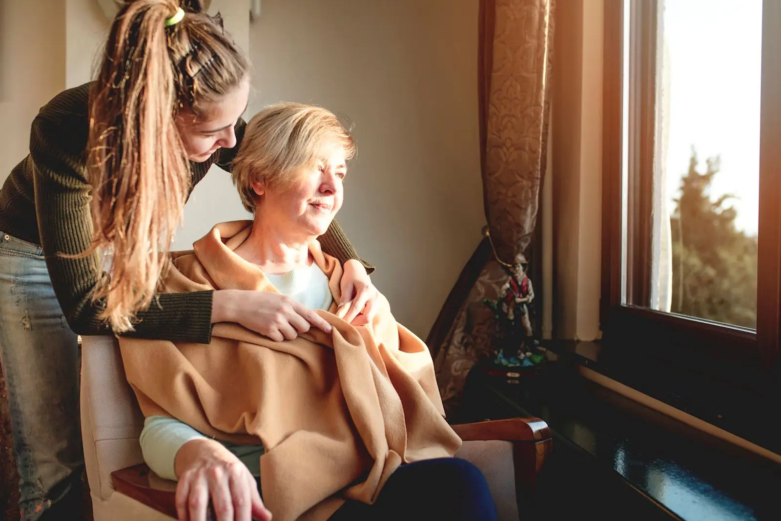 A young woman comforting an older woman sitting in a chair and looking out a sunny window.