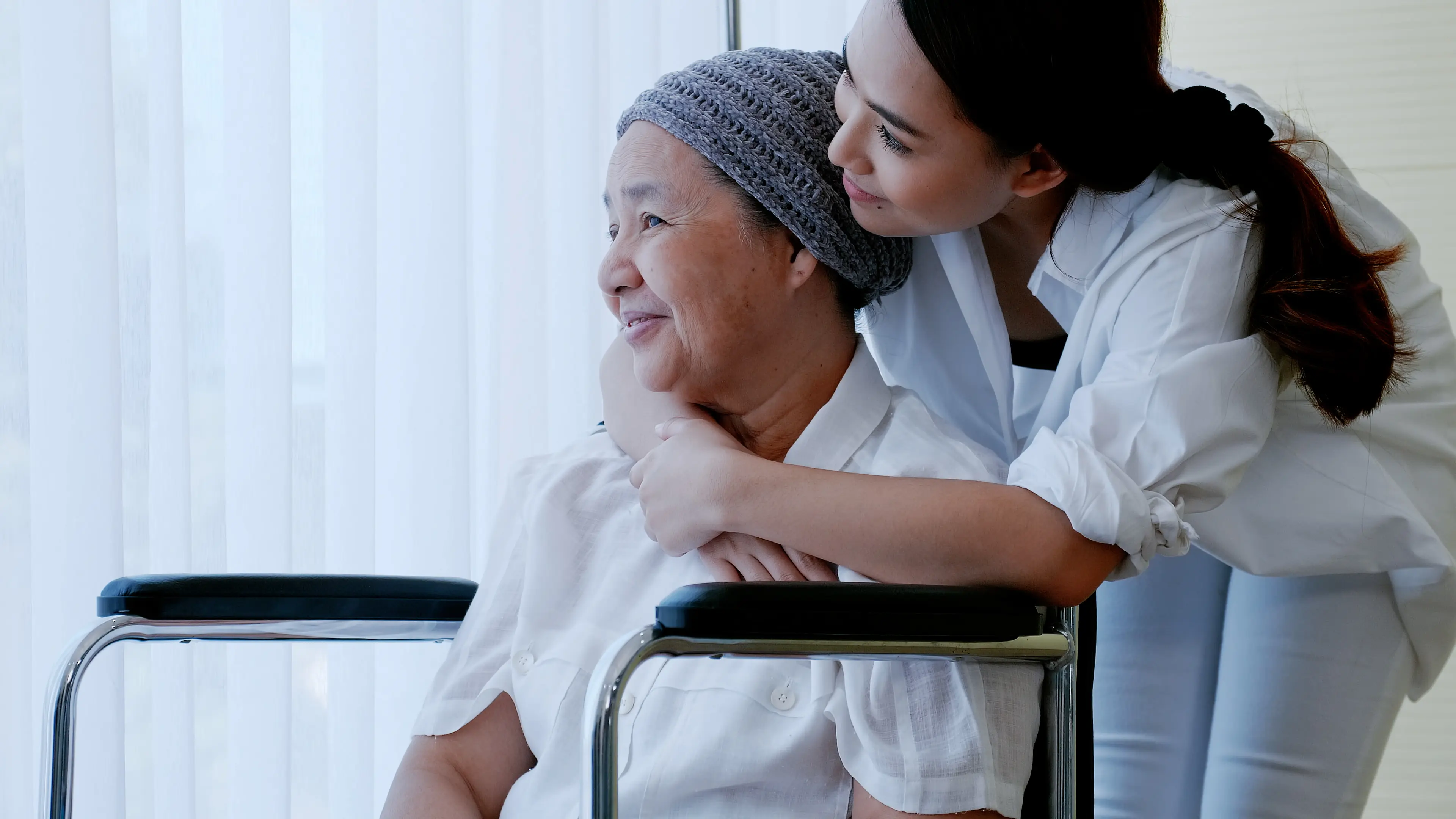 Caregiver hugging an older woman with a head scarf, looking out a window.