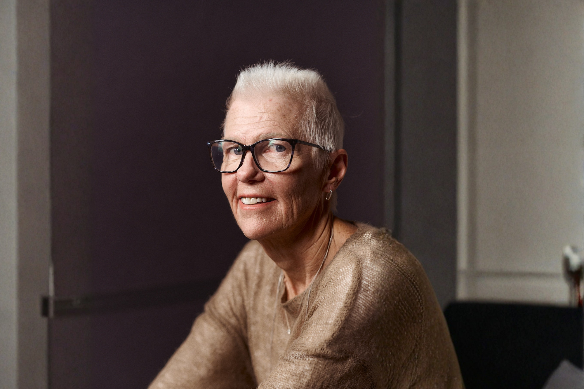 Smiling woman with short white hair and glasses, wearing a gold sweater