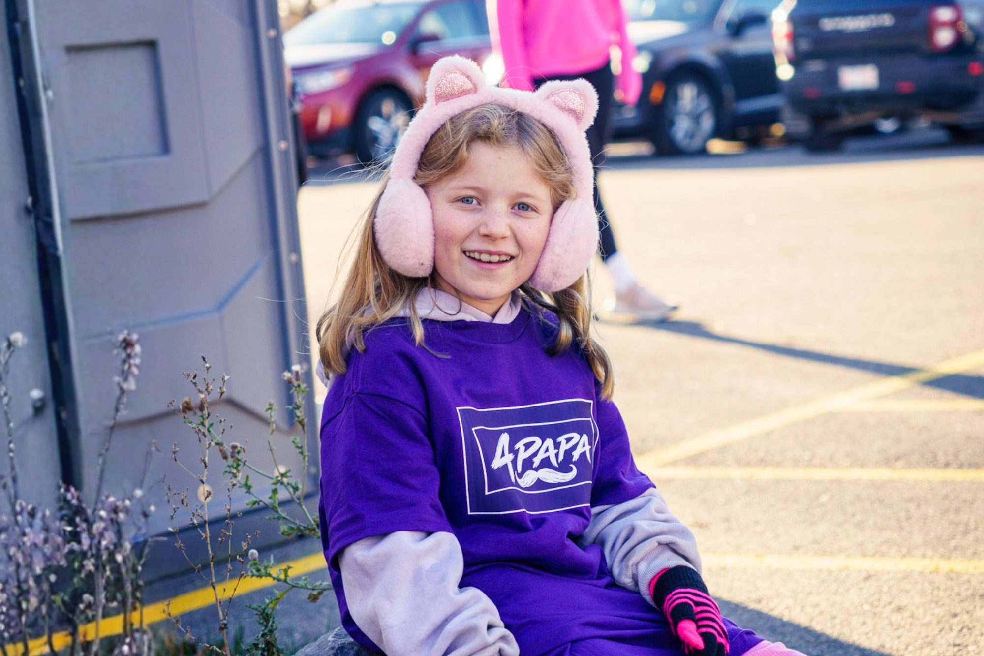Smiling girl in pink cat-ear earmuffs and a purple APAPA T-shirt at an outdoor event