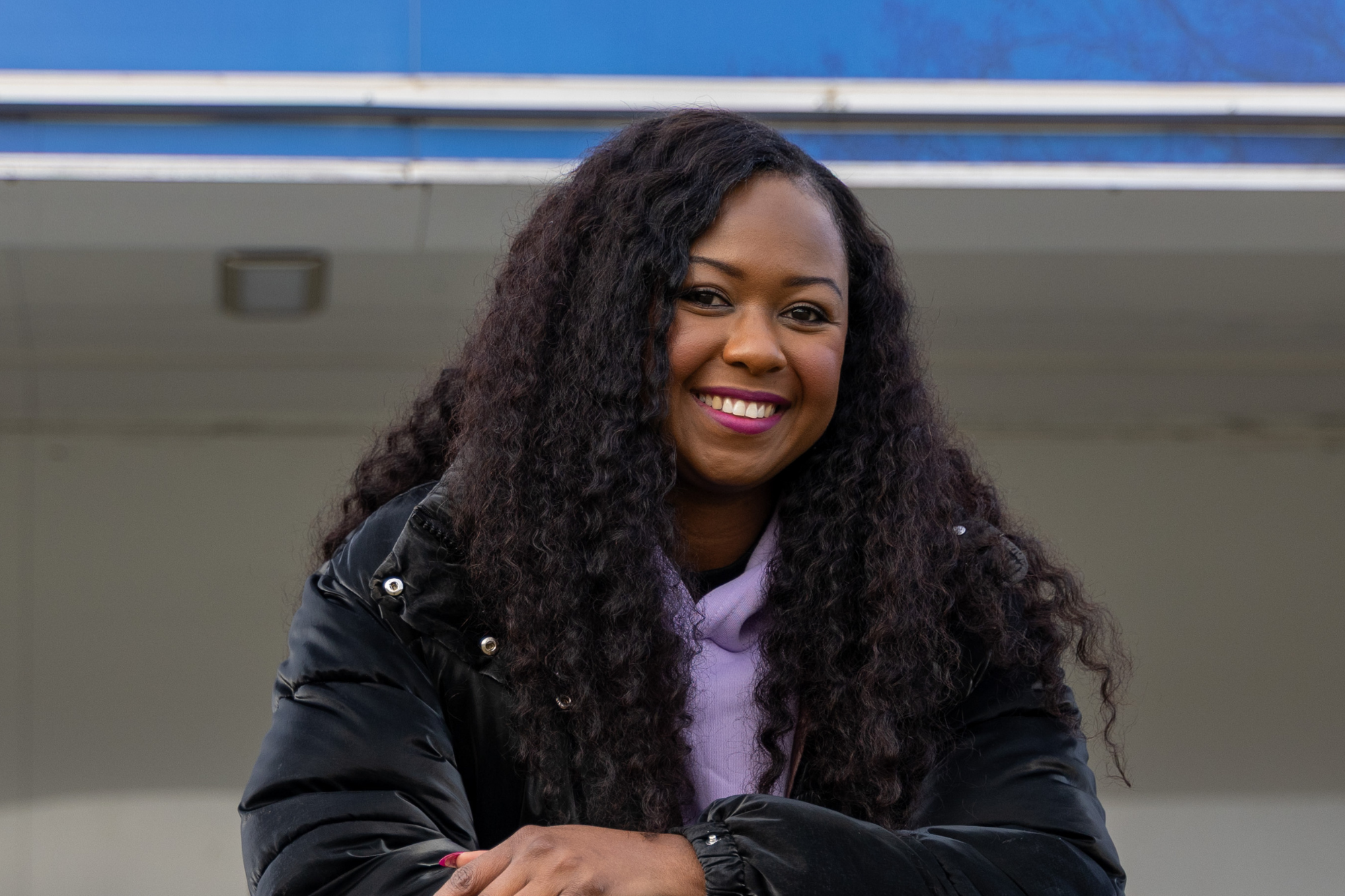 Smiling young woman with long curly hair, wearing a black puffer jacket and lilac hoodie
