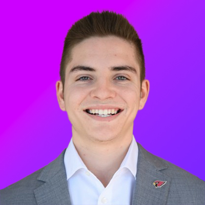 Young man with short dark hair smiling, wearing a gray suit jacket with a white shirt and a small red pin, against a purple gradient background.
