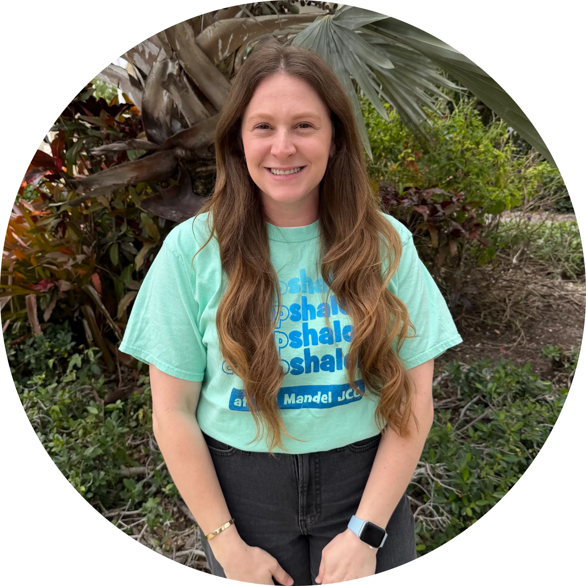 Smiling woman with long wavy brown hair wearing a light green t-shirt and black pants standing outdoors in front of greenery.