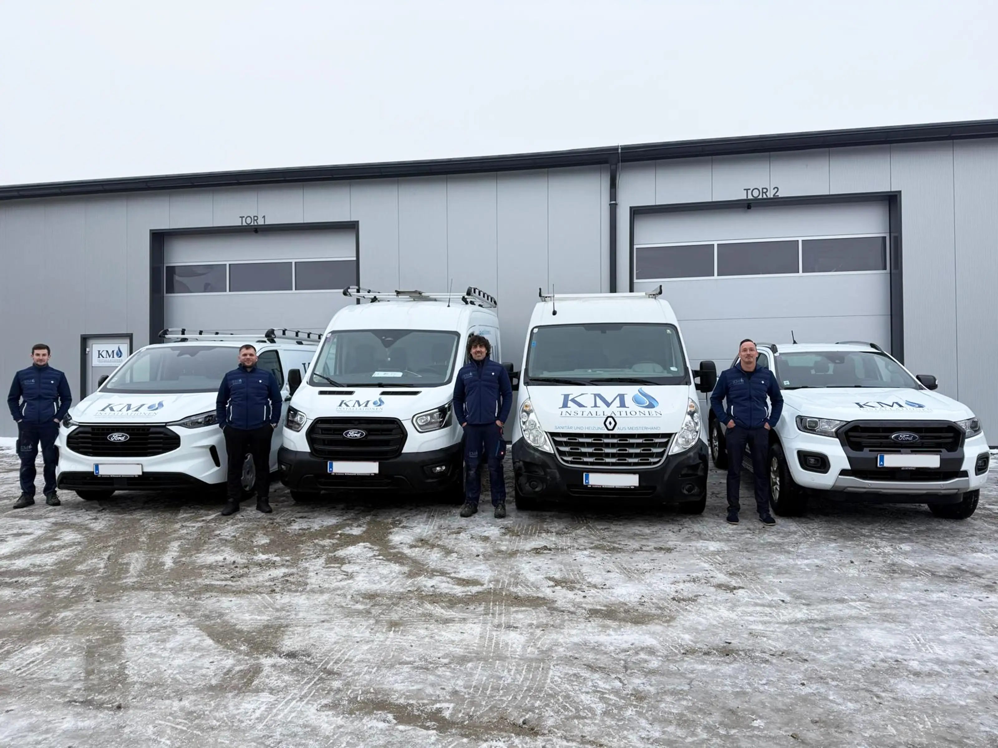 Das Team von KM-Installationen steht aufgereiht neben ihren Autos, vor dem Lager des Betriebs