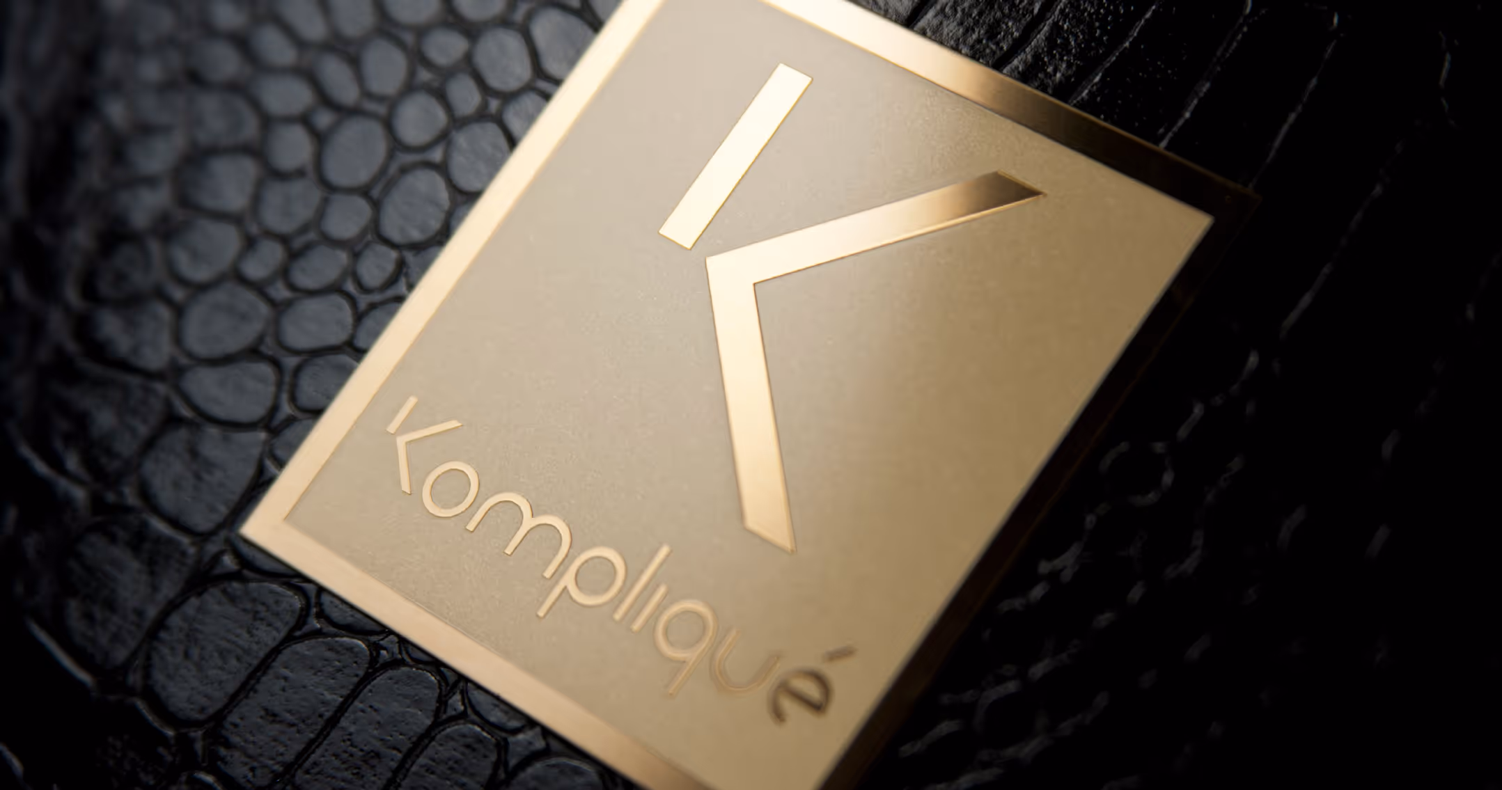 A sleek black box with a gold letter K on top and a dark crocodile skin texture on the right side. It features a small gold plaque engraved with the name mark Kompliqué on the side. The box is set against a dark background.