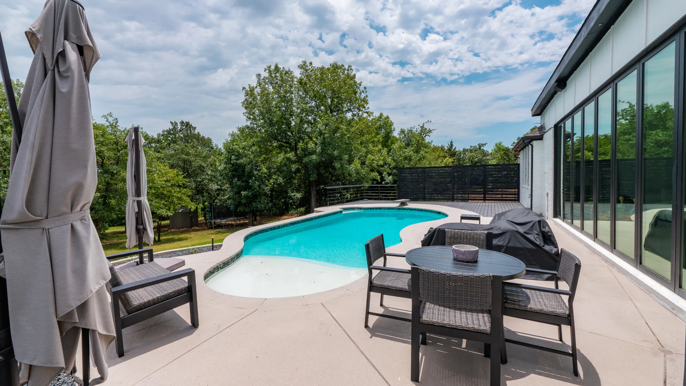 Modern backyard swimming pool with patio seating, umbrellas, and a covered outdoor living area.