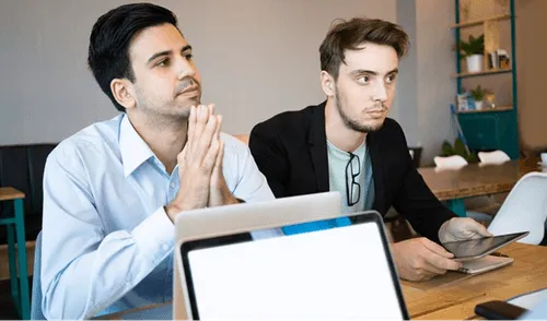 Two men at a meeting table looking attentively at a laptop screen, one holding a smartphone.