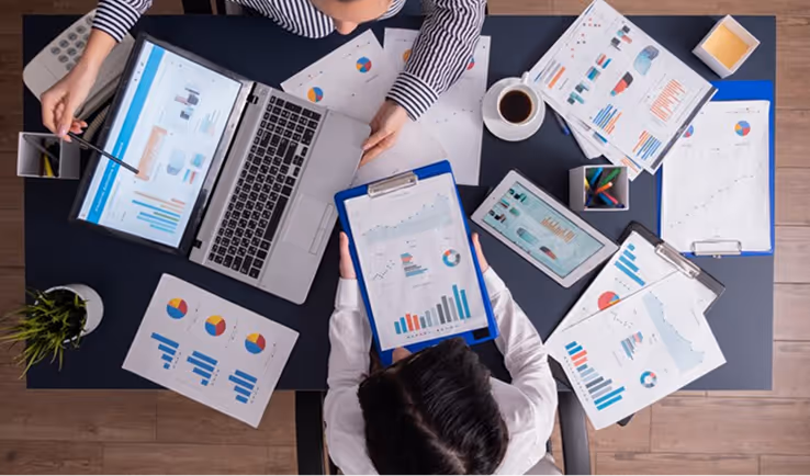 Two people working on a table with a laptop, tablet, and multiple charts and graphs on paper.