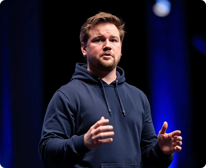 Man with beard in navy hoodie gesturing with hands against dark blue background on stage.