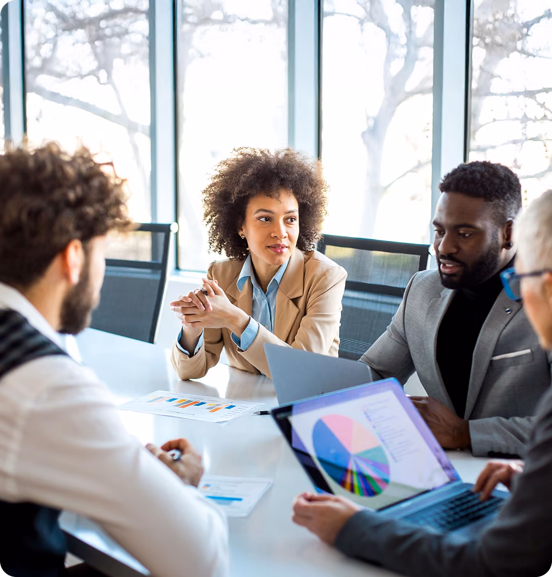 Four diverse professionals in a meeting room discussing data charts from laptops and papers.