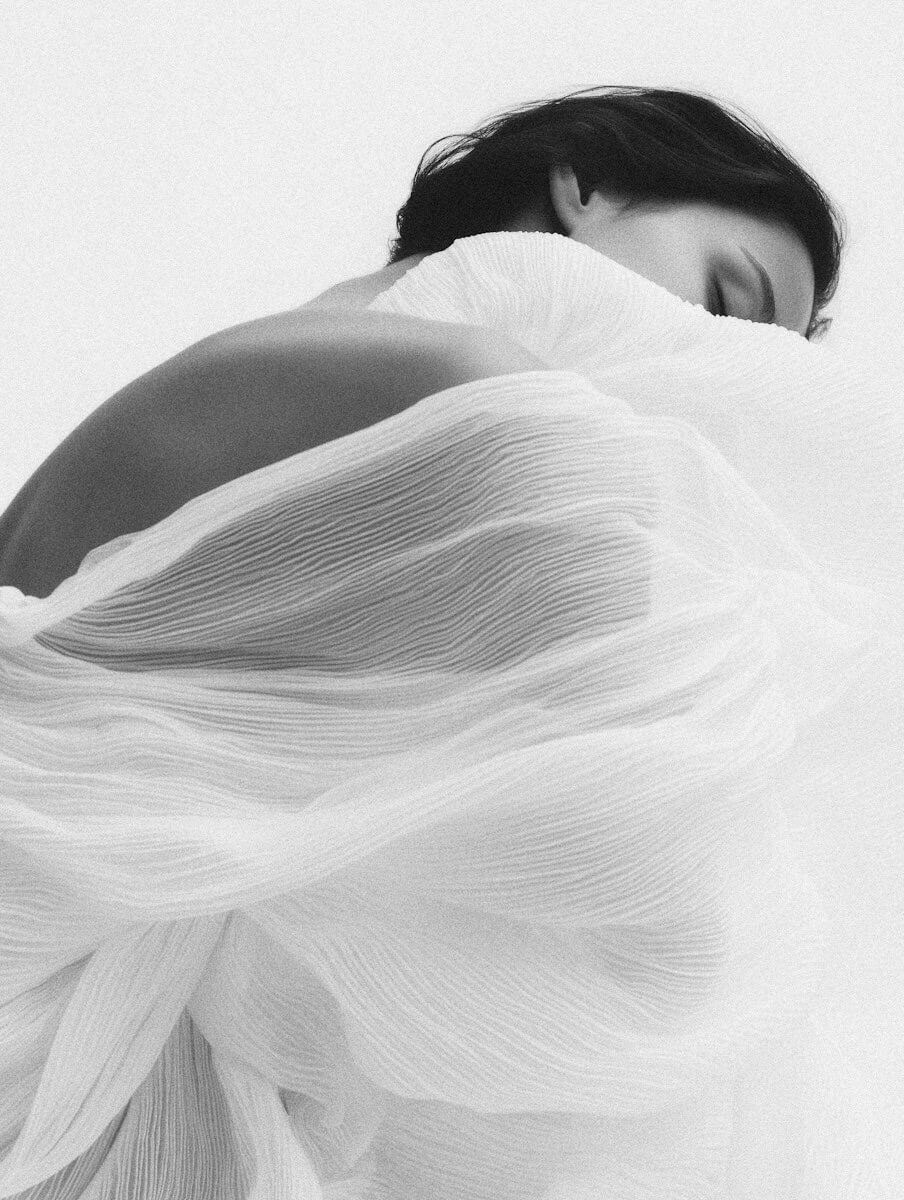Black and white portrait of a woman wrapped in soft, pleated white fabric. Minimalist fashion editorial with ethereal texture.